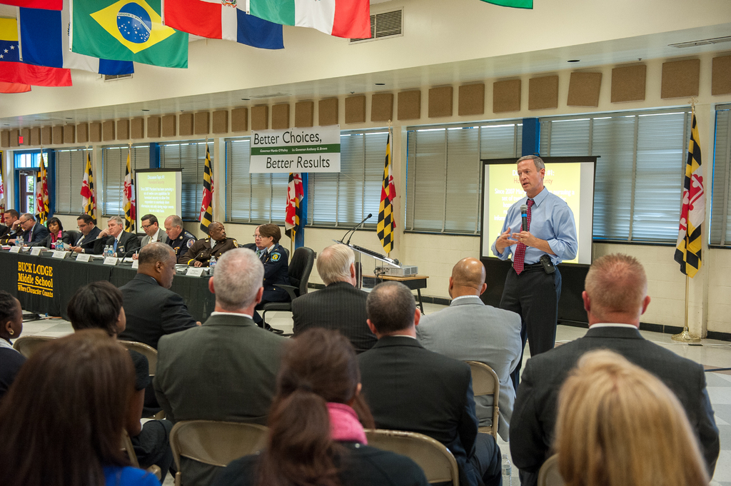 Eine Gruppe von Menschen, die in einem Auditorium auf Stühlen sitzen, wobei eine Person mit einem Handsender vorne steht und in ein Mikrofon spricht. Hinter dem Redner ist ein Projektionsschirm zu sehen, und ein Projektor steht auf einem Tisch rechts daneben. Eine Gruppe von Personen sitzt an einem Tisch mit Wasserflaschen, und Fahnen sind an den Wänden und an der Decke zu sehen. Die Fenster sind abgedeckt, und auf der linken Seite ist eine Tür zu sehen.