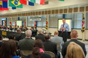 Eine Gruppe von Menschen, die in einem Auditorium auf Stühlen sitzen, wobei eine Person mit einem Handsender vorne steht und in ein Mikrofon spricht. Hinter dem Redner ist ein Projektionsschirm zu sehen, und ein Projektor steht auf einem Tisch rechts daneben. Eine Gruppe von Personen sitzt an einem Tisch mit Wasserflaschen, und Fahnen sind an den Wänden und an der Decke zu sehen. Die Fenster sind abgedeckt, und auf der linken Seite ist eine Tür zu sehen.