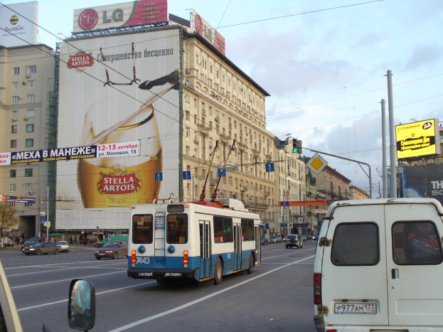 Eine belebte Stadtstraße mit mehreren Fahrzeugen, darunter ein Bus und ein Lieferwagen, umgeben von Gebäuden mit Fenstern und Schildern, Strommasten mit Drähten, Verkehrsampeln und einem bewölkten Himmel.