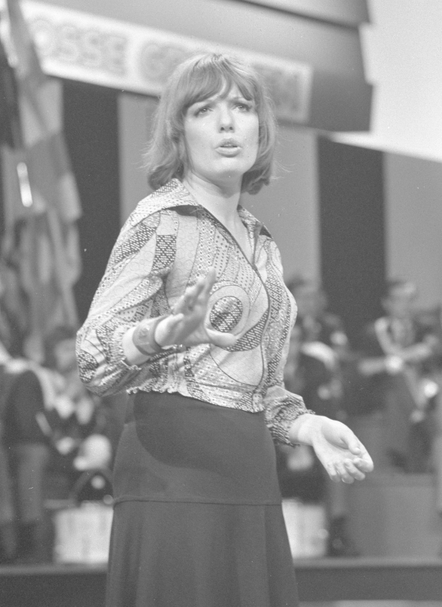 Schwarzes und weißes Foto einer Frau in einem weißen Hemd und schwarzer Hose, die auf einer Bühne steht und einen ernsten Gesichtsausdruck hat, umgeben von einer Gruppe von Menschen und einer Wand im Hintergrund.