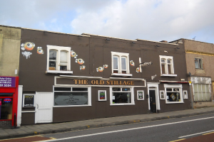 Eine Straßenszene außerhalb der Stadt mit einer Straße, einem Fußweg und Gebäuden mit sichtbaren Türen und Fenstern, einschließlich einer roten Wand und einem violetten Schild mit Text, unter einem klaren Himmel.