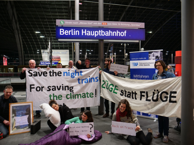 Eine Gruppe von Menschen sitzt auf dem Boden des Berliner Hauptbahnhofs, hält ein Banner mit der Aufschrift "Retten Sie die Nachtzüge zum Schutz des Klimas", umgeben von Kissen, einem Foto Rahmen und anderen Gegenständen, mit Brettern, Säulen, Lampen und Geländern im Hintergrund.