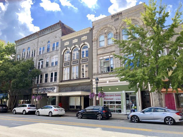 Eine Stadtstraße mit parkenden Autos, eine Mischung aus alten und neuen Gebäuden mit Fenstern, Bäumen, Laternen und einem bewölkten Himmel im Hintergrund.