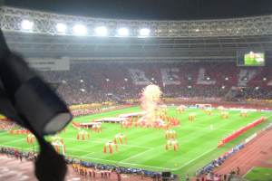 Ein Stadion mit Flutlicht, Eisengittern, Zuschauern, stehenden Personen, Werbetafeln, Anzeigebildschirmen, Überwachungskameras, Fahnen und Dekorationen.