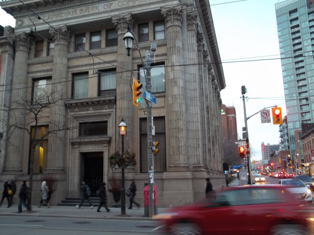 Eine belebte Stadtstraße mit Fahrzeugen, Fußgängern, Gebäuden und Bäumen, mit einem großen Gebäude im Vordergrund und einem roten Auto, das die Straße entlangfährt.