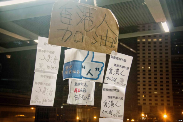 Eine Gruppe von Menschen steht vor einem Gebäude in der Nacht, mit beleuchteten Protestschildern an der Decke.