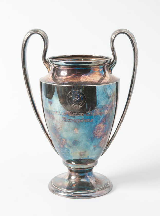 Der UEFA-Champions-League-Pokal, ein stählerner Becher mit zwei Griffen und einem blauen und silbernen Farbschema, der mit 'UEFA Champions League' auf einem weißen Hintergrund beschriftet ist.