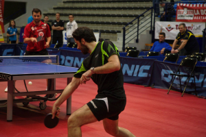 Eine Gruppe von Menschen in verschiedenen Farben, darunter schwarz, weiß und blau, sitzt um einen Tisch mit einem Spiel in einem Raum mit Stühlen und einer Treppe im Hintergrund.