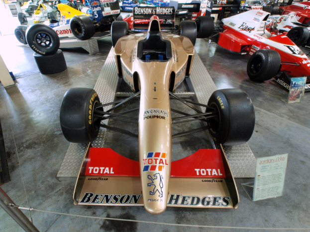 Eine Gruppe von Rennwagen in einem Motorsportmuseum ausgestellt, getrennt durch einen Metallzaun mit informativem Text im Hintergrund.