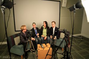 Eine Gruppe von drei Personen sitzt auf einem Sofa und gibt ein Interview, wobei eine Person in einem schwarzen Mantel und Hose links, eine Person in einem Mantel und blauer Krawatte mit einem Mikrofon in der Mitte und eine strahlende Person rechts zu sehen ist. Ein Mikrofonrecorder befindet sich auf dem Tisch in der Mitte, Kameras sind auf der rechten und linken Seite und große Scheinwerfer sind vorhanden, mit einem Projektionsschirm im Hintergrund.