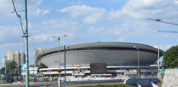 Großes Stadion mit Fahrzeugen, Gebäuden und Grünflächen vorne, unter einem bewölkten Himmel.