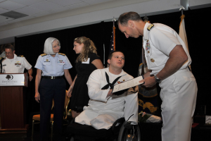 Ein Mann im Rollstuhl erhält eine Auszeichnung von einem stehenden Mann, während andere zusehen, einer hält ein Buch, mit einem Podium, Mikrofon und Texttafel links, Flaggen, einem schwarzen Vorhang und Decke im Hintergrund.