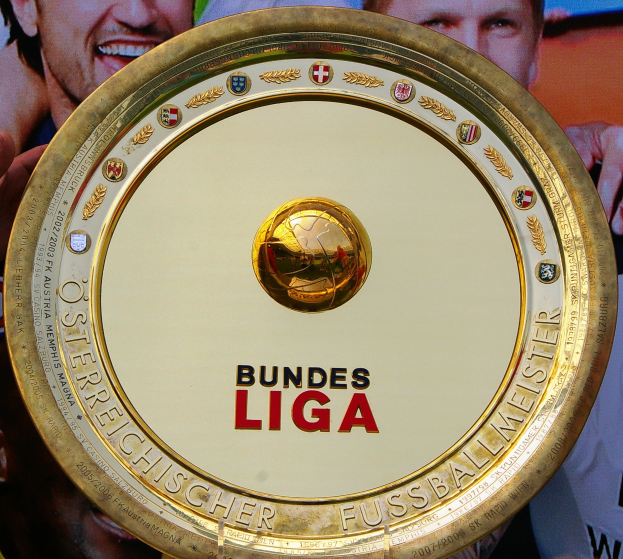 Goldener Schild mit "Bundesliga"-Text in der Hand eines Mannes, mit einem Banner im Hintergrund, das drei Personen zeigt.