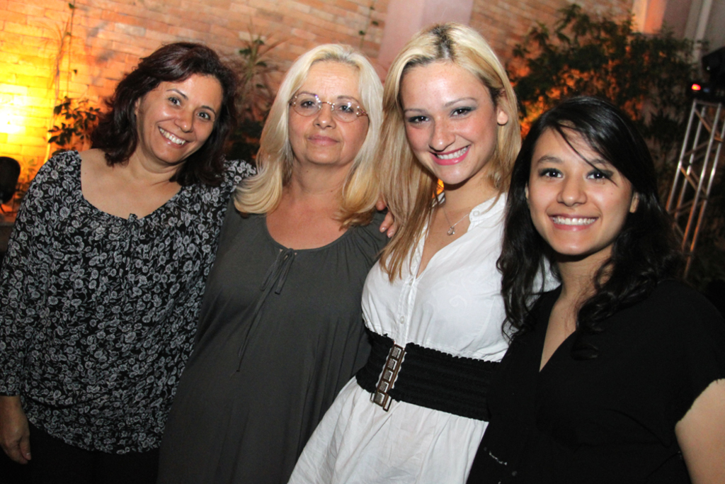 Vier Frauen in der Mitte, die in die Kamera l√§cheln und posen, mit einer Ziegelwand und Pflanzen im Hintergrund.