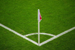 Ein Fußballfeld mit grünem Rasen, weißen Begrenzungslinien und einem weißen Pfosten mit einer roten und weißen Fahne in der Mitte des Strafraums.