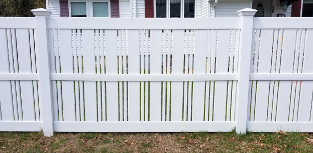Eine weiße Vinyl-Privatsichtzaun steht vor einem Haus mit Fenstern, umgeben von Gras und trockenen Blättern, mit Bäumen im Hintergrund.