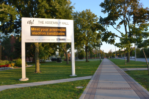 Ein verwittertes Schild auf Gras neben einem Weg und Bäumen zeigt "Die Versammlungshalle - Treffen Sie Ihre Landtagswahlkandidaten" mit einem bewölkten Himmel, Fahrzeugen, einem Gebäude und Straßenlaternen im Hintergrund.