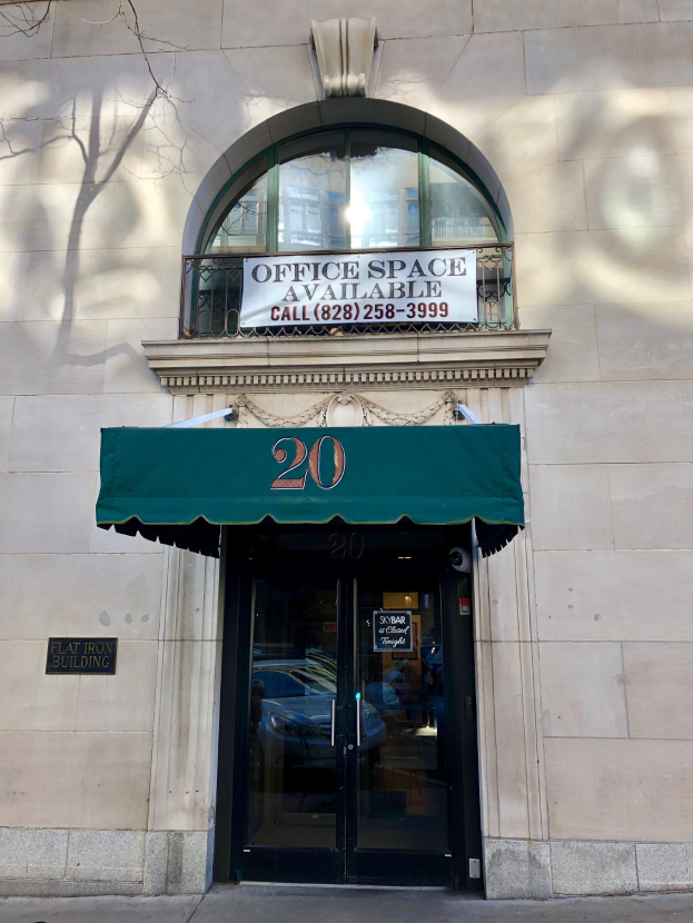 Glasstür mit grüner Markise an einem Gebäude, mit einer Tafel, die "Büroräume zu vermieten" anzeigt, einem Geländer und einem Baum links.