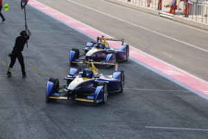 Zwei Formel-E-Autos auf einer Rennstrecke mit einem Flaggenträger zwischen ihnen, Menschen auf der linken Seite und Zuschauern und Gittern im Hintergrund.