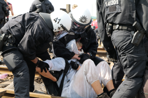 Eine Gruppe von Polizisten in Einsatzausrüstung verhaftet einen Mann in einem weißen Kleid auf einem Bahngleis, mit anderen im Hintergrund sichtbar.