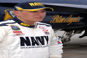 Ein Mann in einem weißen Jackett und blauer Mütze steht vor einem Flugzeug und sieht entschlossen aus.