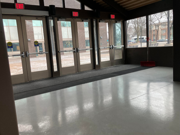 Großer Raum mit hoher Decke, zahlreichen Fenstern und Türen, mit einem Besen auf dem Boden, Schildern mit Text, einem sichtbaren Dach, umliegenden Bäumen, Fahrzeugen auf dem Boden, benachbarten Gebäuden mit Fenstern und einem bewölkten Himmel, was auf einen gewerblichen Bodenreinigungsservice hinweist.