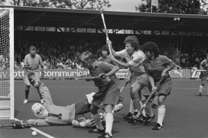 Schwarzes Bild einer Gruppe von Frauen, die Feldhockey spielen, mit Helmen und Stöcken, einem Torpfosten links und Zuschauern im Hintergrund.