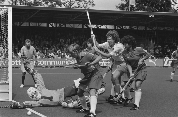 Schwarzes Bild einer Gruppe von Frauen, die Feldhockey spielen, mit Helmen und Stöcken, einem Torpfosten links und Zuschauern im Hintergrund.