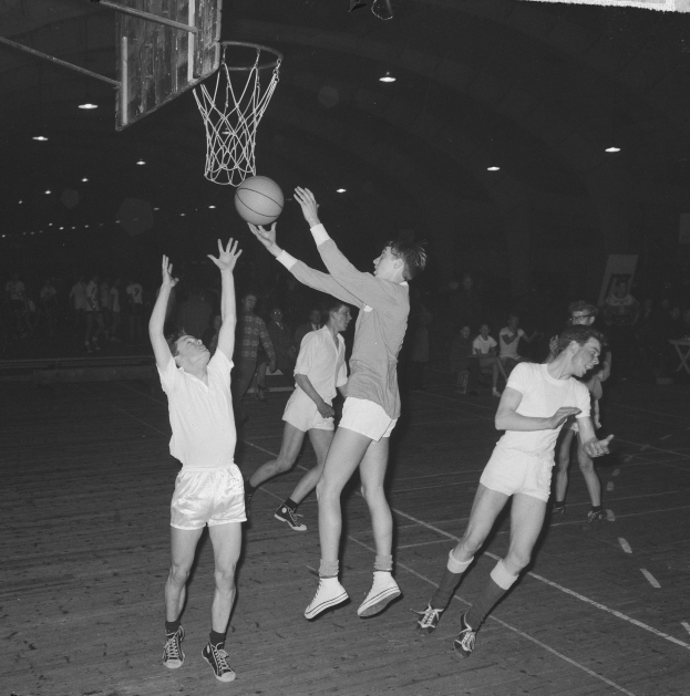Eine Gruppe junger Männer, die Basketball in einer Turnhalle spielen, mit einem Basketballkorb im Hintergrund und Menschen, die sitzen und stehen, in Schwarzweiß dargestellt.