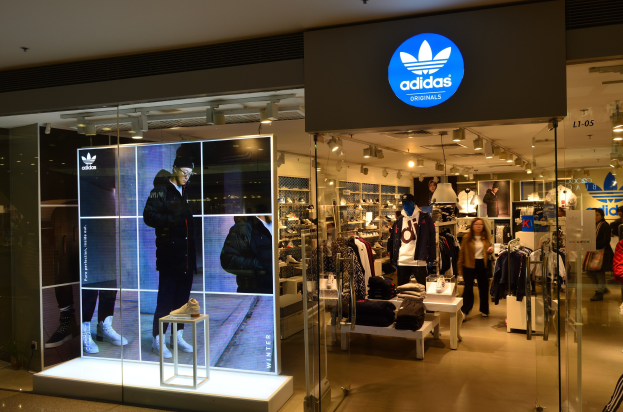 Ein Schaufenster für Adidas Originals in einem Einkaufszentrum mit Schaufensterpuppen, hängenden Kleidern, präsentierten Schuhen und beleuchteter Markenwerbung.