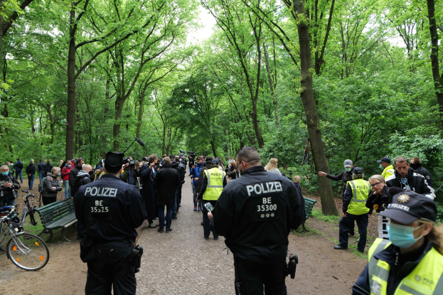 Eine Gruppe von Polizisten steht vor einer Menge von Menschen, einige tragen Mönen und Masken, mit Fahrrädern und einer Bank im Vordergrund und Bäumen und Himmel im Hintergrund, wahrscheinlich bei einer Anti-Terror-Demonstration in Berlin.