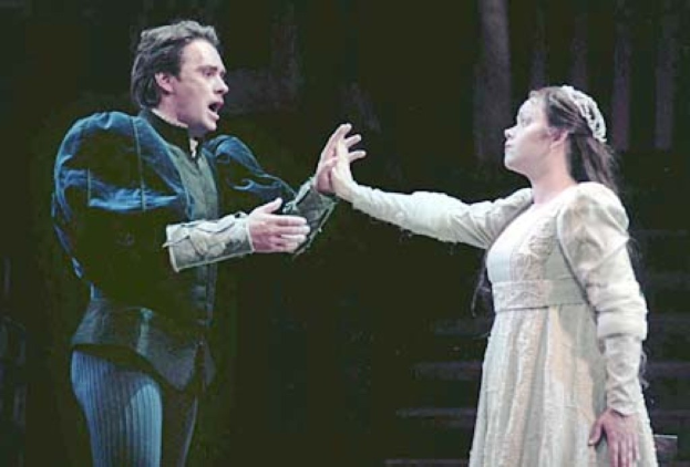 Ein Mann in einem schwarzen Anzug und weißem Hemd und eine Frau in einem langen Kleid stehen auf einer Bühne und sehen ernst aus, wobei der Mann ein Objekt hält und eine dramatische dunkle Kulisse mit einer Treppe im Hintergrund zu sehen ist.