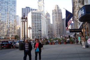 Eine belebte Straßenszene mit Menschen, Blumentöpfen, Autos, Straßenlaternen, Gebäuden, Geschäften und Flaggen auf der rechten Seite.