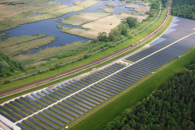 Luftaufnahme einer Solar-Farm mit Panelen auf einer Wiese, umgeben von Baumen und Wasser, mit einem Zug auf einer nahen Bahnschiene.