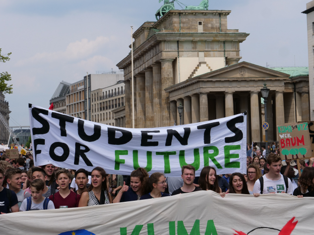 Gruppe von Studenten, die in Berlin marschieren und eine bunt bemalte "Students for Future"-Fahne tragen, vor einem Hintergrund aus Gebäuden, Bäumen und Himmel.