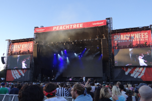 Eine große Menschenmenge vor einer hell erleuchteten Bühne beim Shaky Knees Music Festival, auf der Musiker spielen, mit Bannern und einem Geländer unten und einem klaren blauen Himmel im Hintergrund.