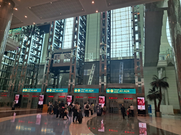 Eine große Gruppe von Menschen geht durch eine Flughafenhalle mit hohen Gebäuden, Bäumen, Texttafeln, Säulen und Deckenleuchten, viele tragen Taschen.