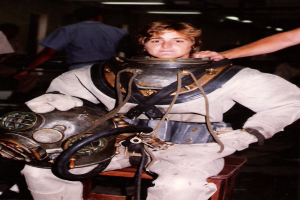 Eine Frau in einem Tauchanzug sitzt auf einem Stuhl und hält einen Tauchhelm, mit einigen Menschen und Lichtern im Hintergrund.