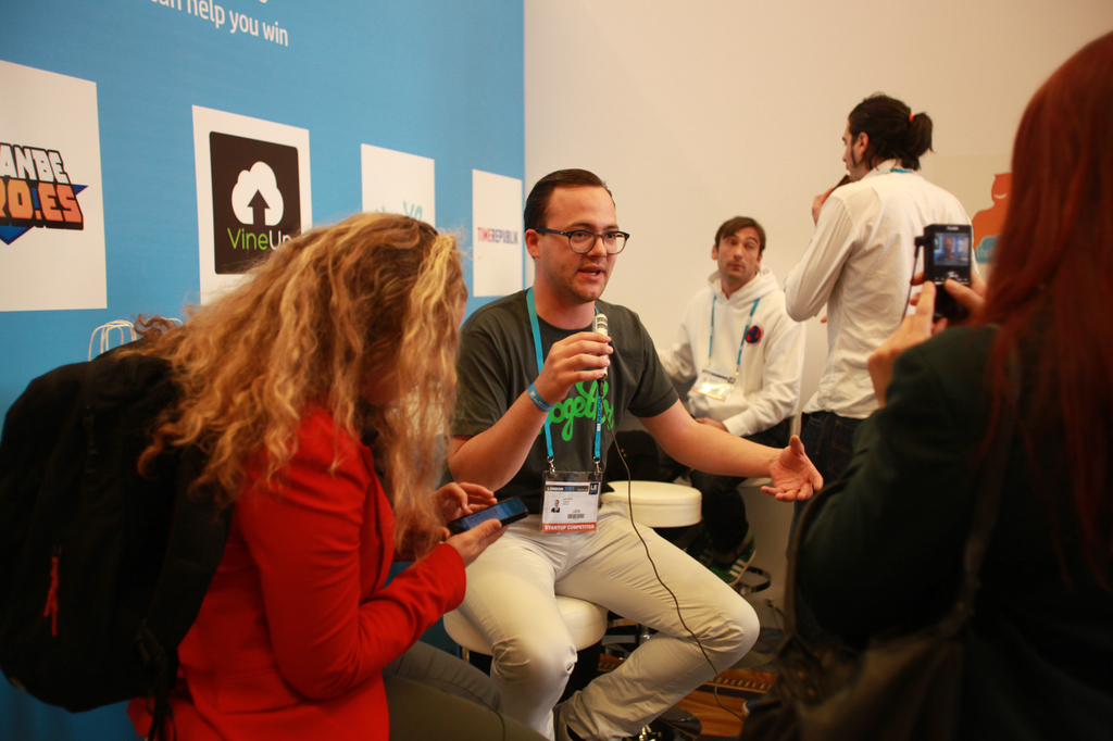 Ein Interview-Szenario mit Menschen mit Mikrofonen und einer Kamera, ein Banner auf der linken Seite und Personen im Vordergrund und in der Mitte, mit einem klaren Hintergrund.