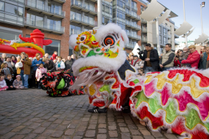 Ein lebendiges chinesisches Neujahrsfest in Amsterdam mit einem Löwen tanzen vor einem Publikum, einige halten Kameras, vor einem Hintergrund von Gebäuden, Laternenmasten und einem klaren blauen Himmel.