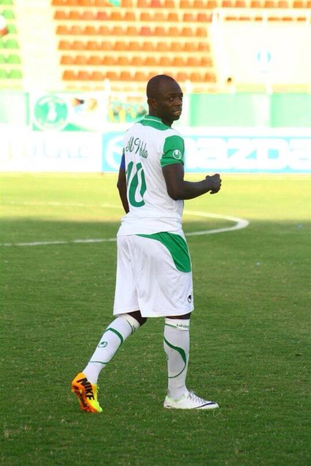 Ein Mann in einem grün-weißen Fußballtrikot feiert ein Tor auf einem Feld, umgeben von Werbetafeln und Stühlen, wahrscheinlich nach dem Sieg seines Teams bei der Afrikameisterschaft.
