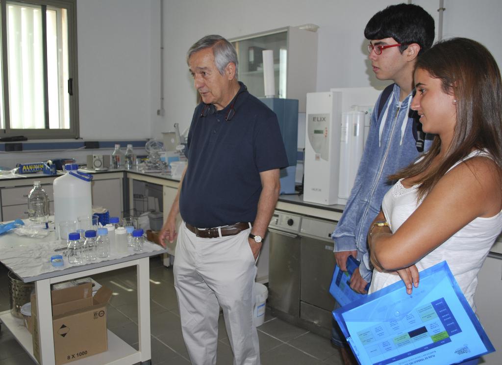 Ein Mann und eine Frau mit Akten stehen neben einem Tisch mit Flaschen, Behältern, Tüchern und einer Kartonschachtel, mit einem elektrischen Gerät, mehr Flaschen und Gegenständen im Hintergrund, einem Schrank, einer Wand und einem Fenster.