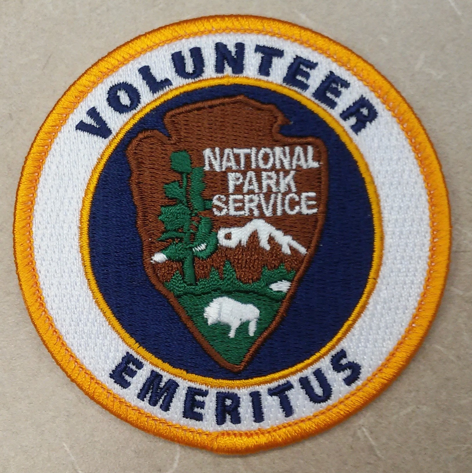 Ein rundes Patch mit blauem Rand und weißem Hintergrund, das in der Mitte schwarze Schrift zeigt, die 'Volunteer National Park Service Emeritus' lautet.