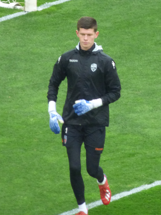 Ein Mann in einer schwarzen Jacke und blauen Handschuhen geht auf einem Fussballfeld mit einem Tor im Hintergrund, trägt Fussballhandschuhe.