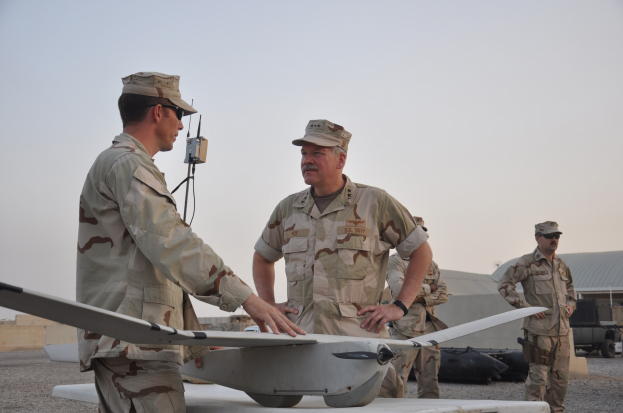 Zwei Männer in Militäruniformen stehen nebeneinander vor einer Drohne, beide tragen Mützen und Schutzbrillen, mit einem Fahrzeug und Himmel im Hintergrund.