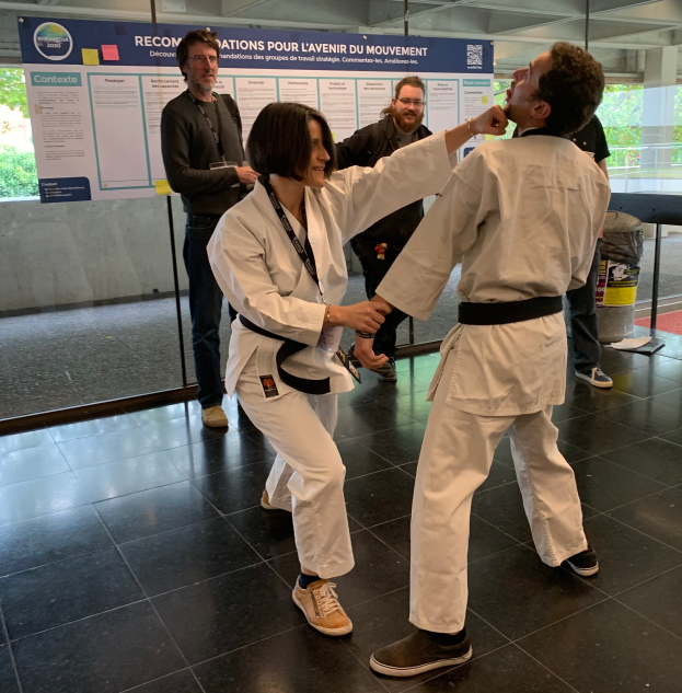 Ein Mann und eine Frau in Karate-Uniformen stehen nebeneinander vor einem Plakat, mit einer Glaswand, die Bäume dahinter zeigt, und einem Mülleimer und einem Tisch mit Gegenständen rechts.