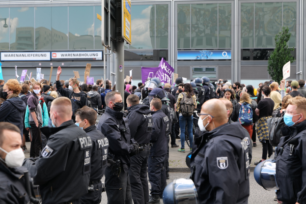 Große Menschenmenge vor einem Gebäude protestierend, einige halten Schilder und tragen Helme, mit einem Schildständer im Vordergrund und einem Baum im Hintergrund.