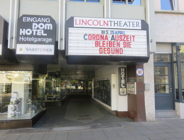 Außenansicht des Lincoln Theaters in Berlin, Deutschland, mit Glasfenstern und -türen und einer Tafel mit Text sowie einem Inneren, das eine belebte Stadtkulisse suggeriert.