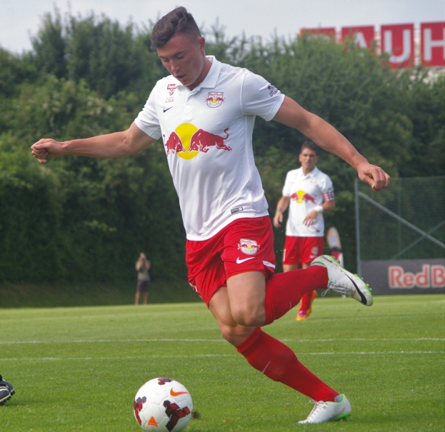 Professioneller Fußballspieler, der einen Ball auf einem Rasenfeld mit dem Schriftzug "RB Leipzig" im Hintergrund unter einem klaren blauen Himmel tritt.