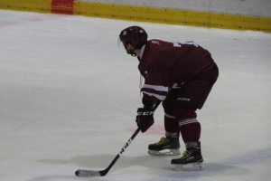Ein Eishockeyspieler auf dem Eis, der einen Stock hält, ein Helm, Handschuhe und Schlittschuhe trägt, mit einer textierten Wand im Hintergrund.